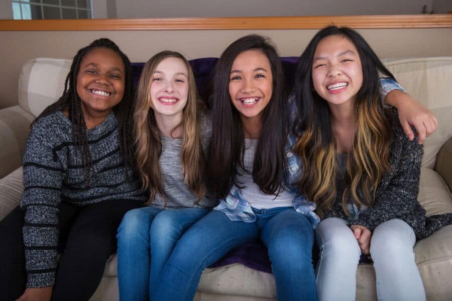 Four smiling children sitting on a couch, arm-in-arm, in a cozy indoor setting. Bright smiles reflect a joyful, friendly atmosphere.