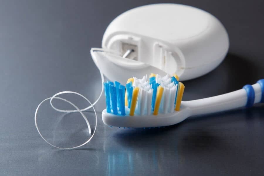 A toothbrush with blue and yellow bristles lies on a gray surface next to an open dental floss container, with floss partially extended.