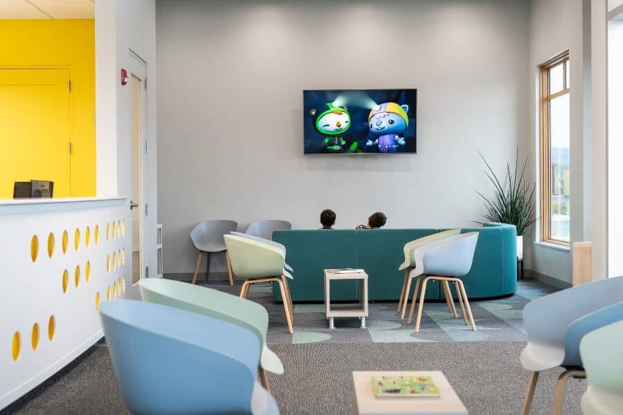 Bright waiting room with modern chairs and playful decor. Two children watch a cartoon on a wall-mounted TV. Yellow accent wall in the background.