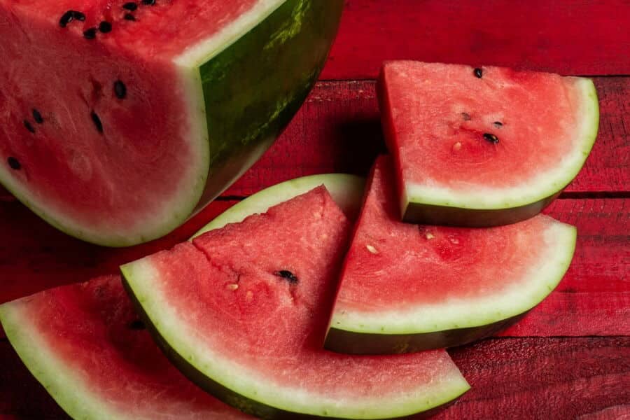 Slices of ripe watermelon with seeds are arranged on a red wooden surface. The juicy texture and vibrant colors are appealing.