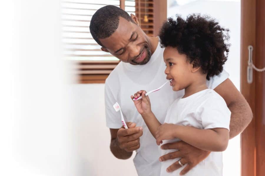 A person and child smiling, brushing teeth together in a bright bathroom. They hold toothbrushes, sharing a moment of hygiene and bonding.