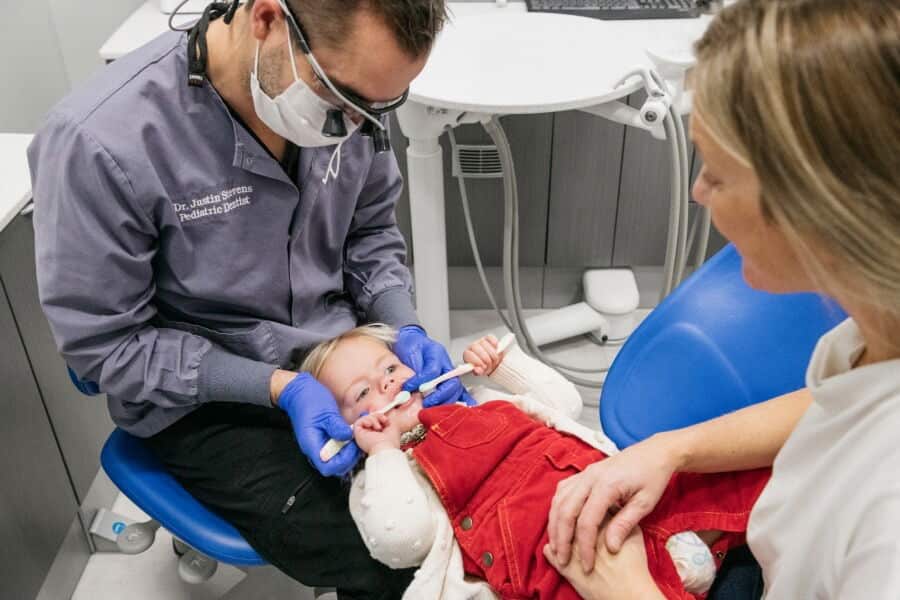 A dentist attends to a child in a dental chair, while another person observes, in a clinical setting with medical equipment.