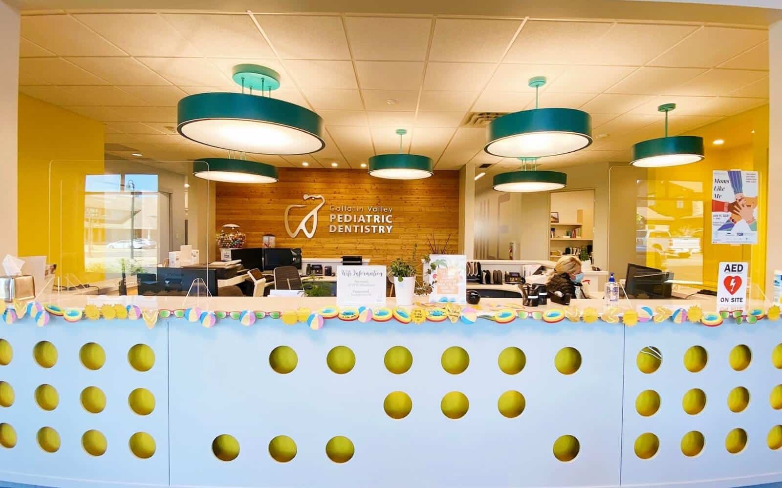 Bright pediatric dentistry office with colorful decor, circular lights, and welcoming reception desk. Staff attending to patients behind the counter. No recognizable landmarks visible.