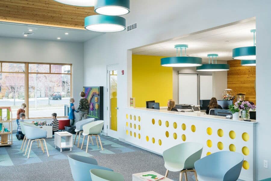 Bright, modern lobby area with colorful furniture. Three people seated at a yellow-accented reception desk. Children playing near a window with toys.