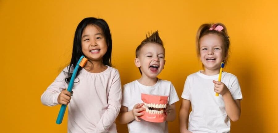 Three children smiling against a yellow background, holding oversized toothbrushes and a dental model, radiating a playful, joyful energy.