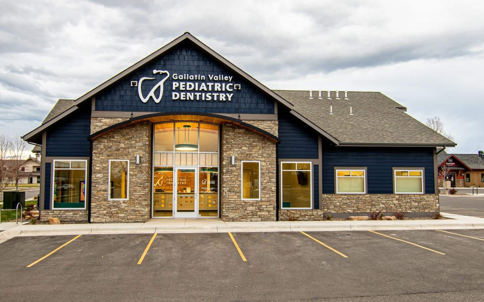 Modern building with "Gallatin Valley Pediatric Dentistry" sign, featuring stone and blue siding, large windows, and a parking area under a cloudy sky.