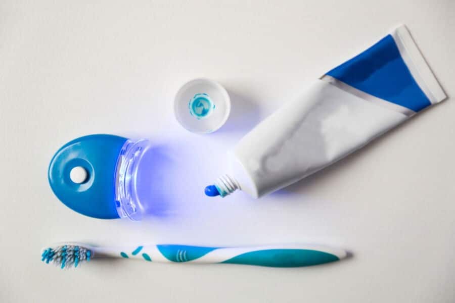 A toothbrush, toothpaste tube, and blue-illuminated dental device are arranged on a white surface, suggesting a dental care setup.