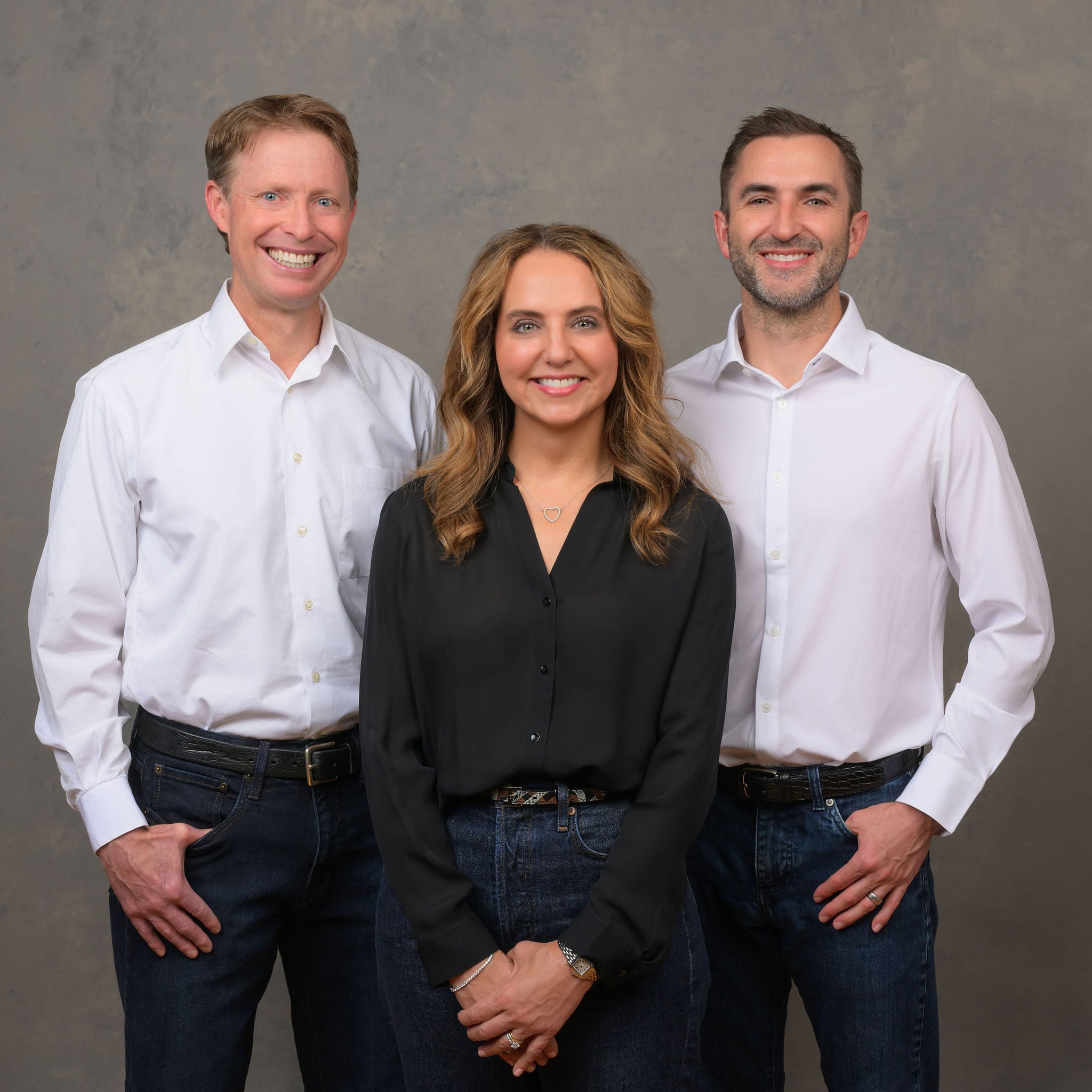Three people are standing together against a plain background, all wearing casual yet professional attire. They're smiling, creating a friendly atmosphere.